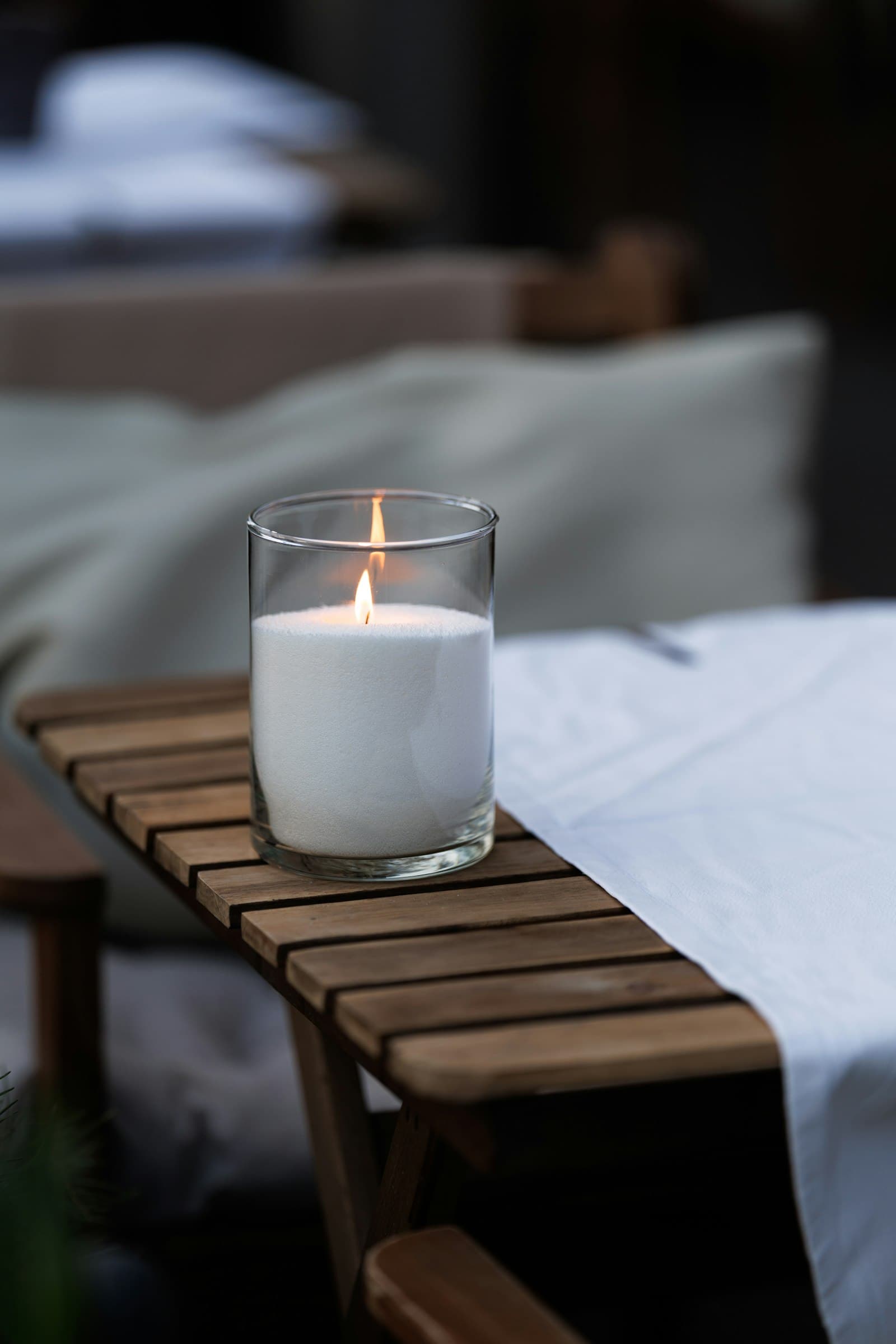 A single lit candle glowing on a wooden bench beside white linen