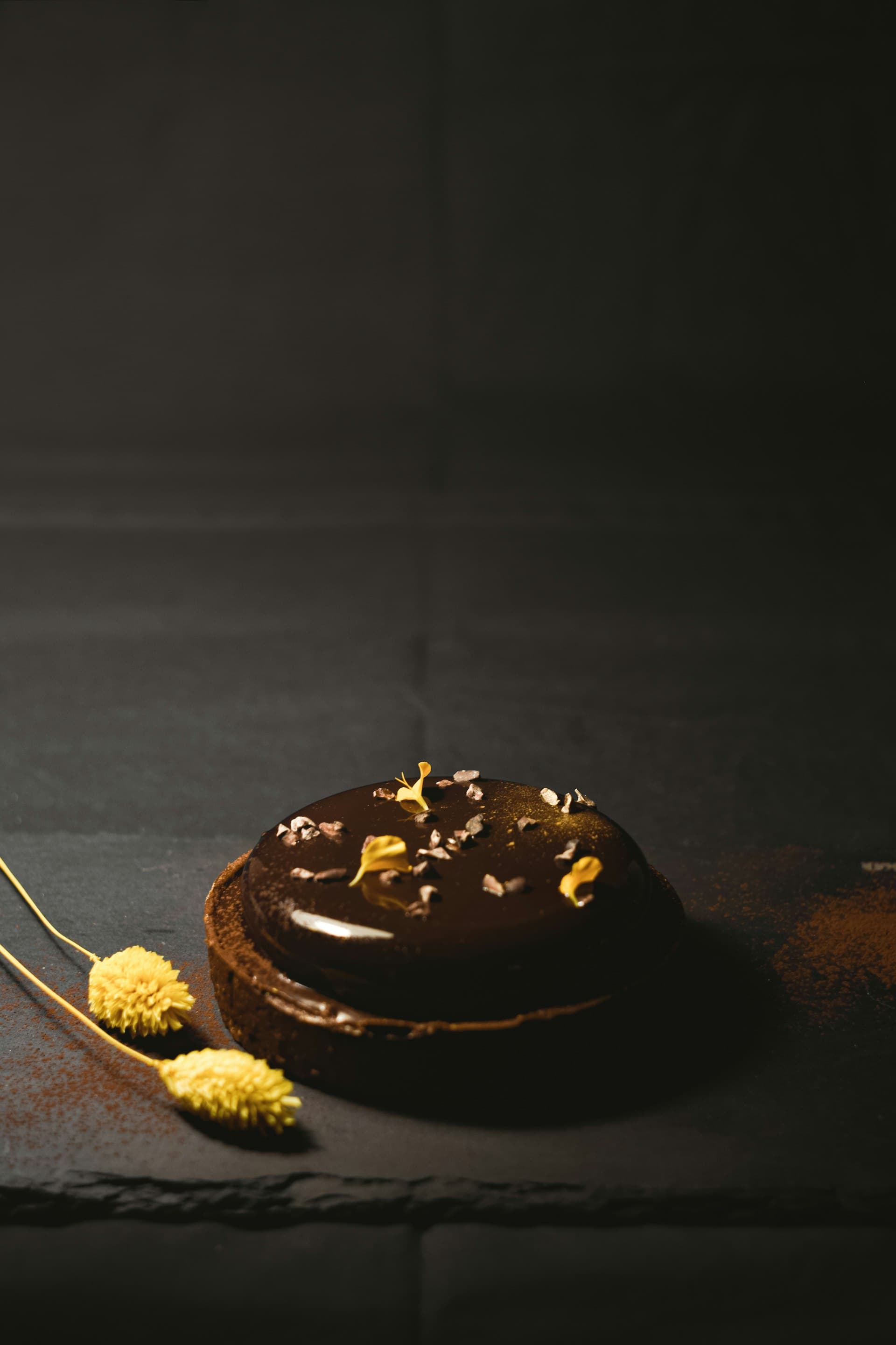 A tall layered chocolate mousse cake dusted with cacao nibs and gold leaf, shot against a black background with a single dramatic light