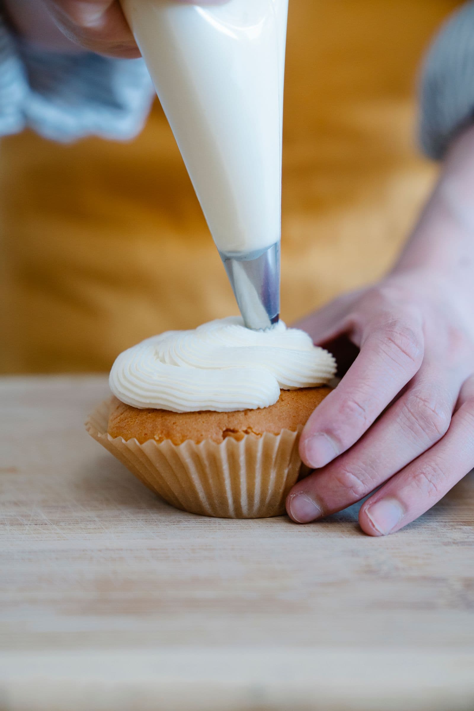 A hand piping a swirl of buttercream onto a cupcake, close-up with warm wood tones