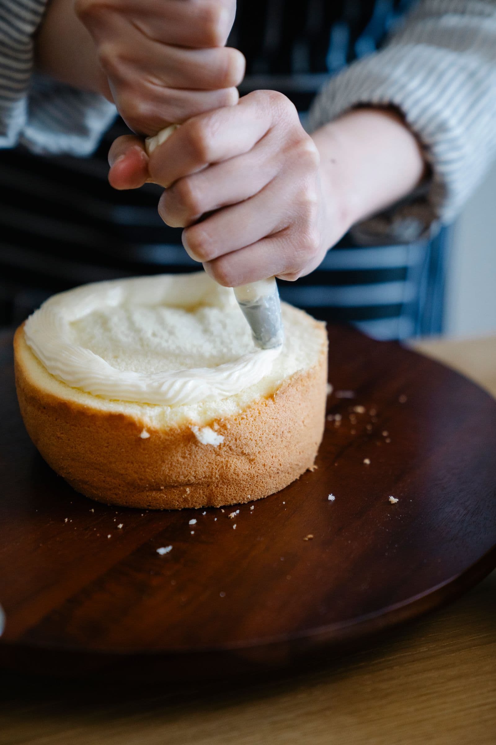 Hands piping whipped cream into a halved sponge cake on a dark wooden turntable, close-up